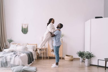 Afrika kökenli Amerikalı erkek, yatağın yanında duruyor ve kadının ellerini kaldırıyor ve dekore edilmiş yatak odasında birbirine bakıyor.