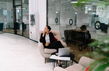 Resmi kıyafetli düşünceli bir kadının modern şirkette cam duvarın yanında cep telefonuyla çalışırken dizüstü bilgisayarla işten ayrılması.