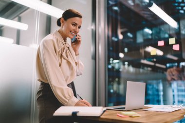 Zarif kıyafetli, gülümseyen genç bir kadın cep telefonuyla konuşuyor. Günlüğüne, modern ofiste dizüstü bilgisayarla notlar yazıyor.