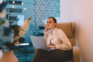 Resmi kıyafetler ve gözlüklerle genç bayan yönetici koltukta oturuyor ve iş yerindeki cam pencerenin yanındaki dizüstü bilgisayarla proje üzerinde çalışıyor.