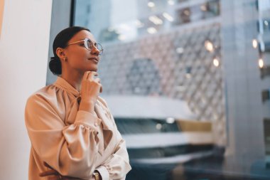 Günümüz modern binasının cam penceresinin yanında gözleri kapalı, parmakları çeneye değen ve gülümseyen gündelik gözlüklü genç bir kadının yan görüntüsü.