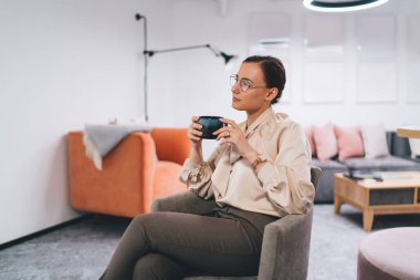Günlük kıyafetleri ve gözlükleriyle koltukta oturan ve elinde bir fincan kahveyle ofis resepsiyonunda dinlenen genç bayan çalışanın yan görüntüsü.