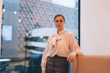 Modern çalışma ortamında panoramik pencereye karşı kameraya bakan şık günlük kıyafetler ve gözlüklü kadın yönetici.