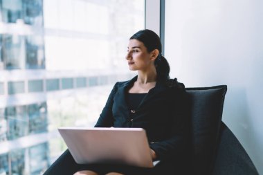 Düşünceli genç bayan serbest çalışan resmi kıyafetlerle Netbook 'a göz atıyor. Kanepede oturmuş, iş yerindeki pencereye bakarak proje üzerinde çalışıyor.