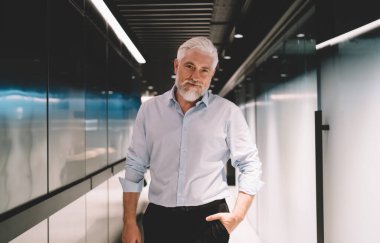 Zarif gömlekli yakışıklı yaşlı adam modern koridorun cam duvarının yanında dururken kameraya bakıyor ve el ele tutuşuyor.