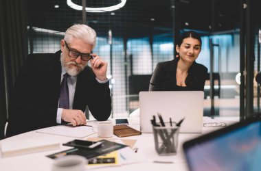 Resmi giyimde konsantre başarılı iş ortakları dizüstü bilgisayarla masada oturup çağdaş ofiste proje üzerinde birlikte çalışırlar.