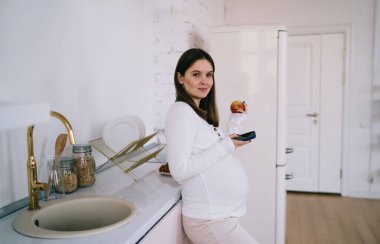 Beyaz arabalı genç bir kadın açık mutfakta duruyor. Elma yiyor. Cep telefonlarını karıştırıyor.