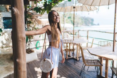 Kendine güvenen genç bayan milenyumu. Uzun saçlı, yaz kıyafetleri içinde. Bayramlarda tropikal kafe verandasında durup kameraya gülümseyen ve bakan.