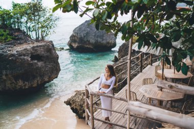Terasta ahşap çitlerin yanında duran ve yaz tatilinin keyfini çıkarırken deniz suyuna bakan yüksek açılı bir kadın.