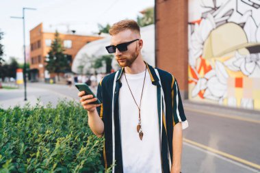 Sıradan giyinmiş genç sakallı bir adam cep telefonuyla mesajlaşırken arka planda bulanık bir şekilde şehrin kaldırımında dikiliyordu.