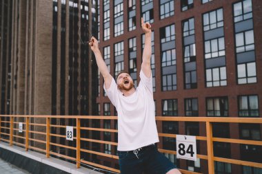 Şehirde modern çok katlı binaların yanında durup başarıyı kutlarken sıradan kıyafetleriyle bağıran ve kollarını kaldıran heyecanlı genç sakallı adam.