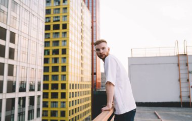 Düşünceli kızıl sakallı bir adamın çatıda dikilip korkuluklara yaslanarak modern çok katlı evler karşısında kameraya bakması.