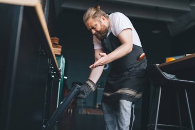 Evdeki mutfakta yemek pişirirken önlüklü, kaşlarını çatan ve sobayı işaret eden kızgın ve cüsseli adamdan.