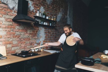 Heyecanlı ve önlüklü adam yuvarlak kesim tahtasıyla savunmada ve evde eğlenirken tahta raptiyeyle tencereye saldırıyor.