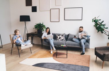 Anne ve babasının cep telefonlarını kullanırken anne bilgisayarını karıştırıyor. Modern oturma odasındaki koltukta boş çerçevelerle oturuyor.
