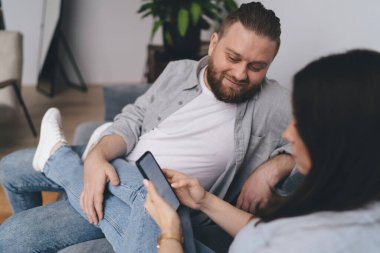 Gülümseyen erkek, kanepede dinlenirken ve evdeki oturma odasında birlikte vakit geçirirken kadının ellerinde cep telefonu ekranına bakıyor.