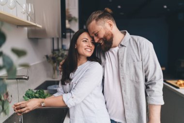 Memnuniyet veren adam mutfakta durup olumlu karısına nazikçe sarılırken bir yandan da kadın musluktan gelen suyla sebze yıkıyor.