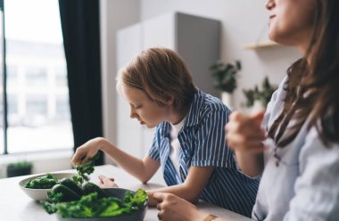 Normal tişörtlü, kadınların yanında duran ve hafta sonu mutfak tezgahında sağlıklı sebze salatası pişiren çocuk.