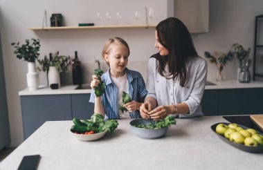 Evde mutfak tezgahında öğle yemeği için sağlıklı salata hazırlarken günlük kıyafetlerle konuşan pozitif anne ve kız.