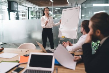 İş kadınlarının konferans odasında iş görüşmeleri sırasında grafikte grafiği açıklarken ahşap masada dizüstü bilgisayarla ve belgelerle oturan resmi kıyafetlerle odaklanmış meslektaşlar.