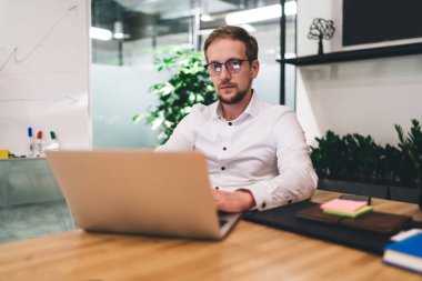 Konsantre sakallı bir adam, gözlüklü, ahşap masada, bilgisayarın yanında oturuyor ve modern ofisteki kameralara bakarken proje üzerinde çalışıyor.