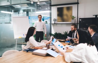 Resmi kıyafet giymiş bir grup iş arkadaşı, geniş konferans odasındaki toplantı sırasında iş arkadaşının sunumunu dinlerken dizüstü bilgisayarları ve belgeleriyle ahşap masada oturuyorlar.