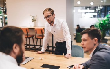 Bir grup odaklanmış iş adamı resmi kıyafetlerini giyip modern çalışma alanlarında iş birliği yaparken proje detaylarını tartışıyor ve birbirlerine bakıyorlar.