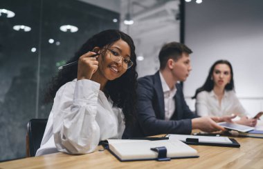 Resmi giyinmiş, gözlüklü, gülümseyen genç siyahi bayan yönetici çeşitli meslektaşlarla masaya oturuyor ve toplantı odasında iş projesi üzerinde çalışıyor.