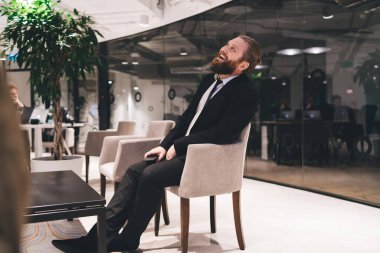 Modern iş merkezinde tatilde koltukta otururken resmi giyinmiş, gülümseyen pozitif sakallı adamın yan görüntüsü.