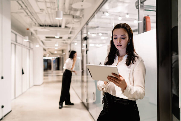 Positive young woman standing with tablet near glass wall of modern office and looking at screen while colleague using smartphone in hallway