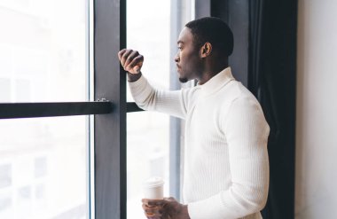 Dakik Afrikalı Afrikalı Amerikalı bir erkek pencerenin yanında durup sıcak aromalı içecek içerken zamanı kontrol ediyor.