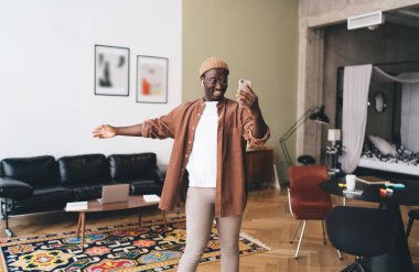 Kulaklık takmış, gülümseyen ve cep telefonu ekranına bakan neşeli Afro-Amerikan erkek. Video görüşmesinde arkadaşlarıyla konuşurken modern oturma odasında elini geriye doğru uzatıyordu.