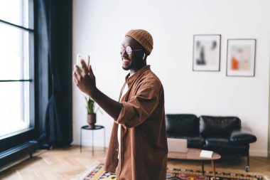 Kulaklık takmış, video görüşmesi sırasında cep telefonu ekranına bakan ve evdeki rahat oturma odasının penceresinin yanında duran kulaklıklı, gülümseyen Afrikalı Amerikalı bir erkek.