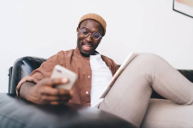 Gözlüklü afro-amerikan genç bir erkek. Evraklar koltukta dinlenirken cep telefonlarını karıştırıyor ve evdeki oturma odasında vakit geçirirken ekrana bakıyor.