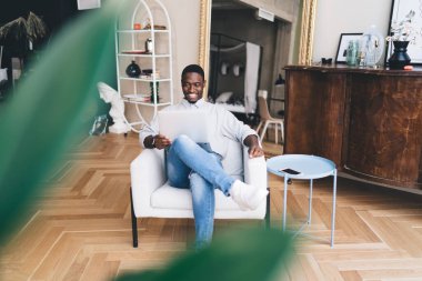 Bulanık yeşil yaprakların arasından Afro-Amerikalı erkek blogcu akıllı telefonlu masanın yanındaki koltukta bacak bacak bacak üstüne atarak rahatlıyor ve evde çevrimiçi blog okurken bilgisayarın ekranına bakıyor.