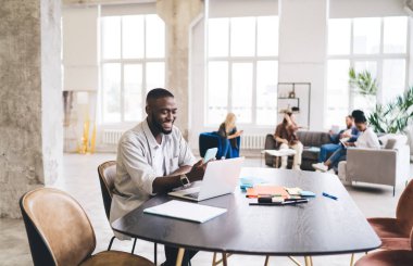 Modern ofisteki dizüstü bilgisayarla masada oturup iş projesi üzerinde çalışırken günlük giysiler içinde cep telefonu kullanan ve gülümseyen siyahi erkek girişimcilerin yan görüntüsü