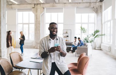 Günlük kıyafetlerle gülümseyen Afrikalı Amerikalı bir adam masa başında oturmuş, cep telefonuyla internette sörf yapıyor ve mesai saatlerinde tavan arası tarzı ofisindeki iş arkadaşlarıyla kameraya bakıyor.