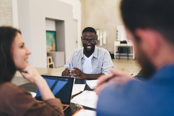 Smiling African American man in glasses taking part in discussion of creative design project while having meeting with blurred partners in light office