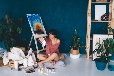 Tasasız genç bayan sanatçı resim sehpası yakınında akıllı telefon kamerası için poz veriyor ve blogunda hobisini gösteren resimler yapıyor. Yaratıcı gülümseyen esmer kadın, yağlı boya resminin yanında kendini fotoğraflıyor. 