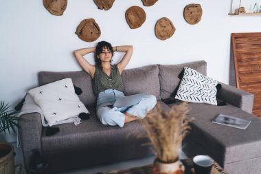 Koyu kıvırcık saçlı, rahat koltukta oturan, bacak bacak üstüne atmış, başı duvara yaslanmış, tahta testereyle süslenmiş genç bir kadın.