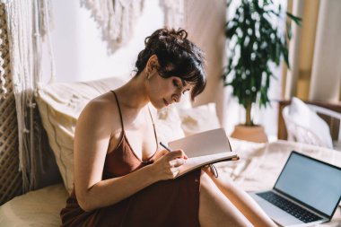 Yan görünüm odaklı İspanyol kadın bilgisayarın yanında oturuyor ve uzak bir proje üzerinde çalışırken not alıyor.