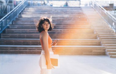 Akıllı telefon ve netbook ile ahşap merdivenlerin yanında duran genç bir kadının kentteki parlak güneş ışığına bakarken yan görüntüsü.