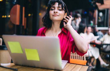 Kendine güvenen bayan girişimci masada oturup saçlarına dokunurken iş projesinde sokak kafesinde dizüstü bilgisayar kullanıyor.