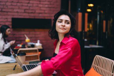 Serbest giyinmiş genç bir bayan netbook ile masada oturuyor ve uzaktan çalışırken mutlu bir şekilde gülümsüyor.