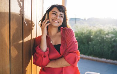 Kırmızı günlük giysiler içinde pozitif genç bir kadın gülümseyip, gözlerini kaçırırken cep telefonuyla konuşuyor ahşap duvara yaslanıyor ve arka planda bulanıklık var.