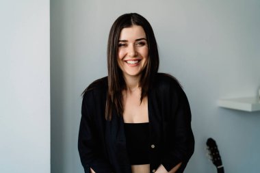 Olumlu genç bayan gitarist gülümseyip kameraya bakarken gitarın yanında beyaz arka planda oturuyor ve boş zamanların tadını çıkarıyor.