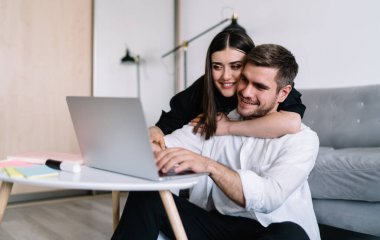 Olumlu genç sakallı erkek arkadaş ve günlük kıyafetlerle gülümseyen kız arkadaş masanın yanında oturuyor ve açık oturma odasındaki dizüstü bilgisayarın klavyesinde yazı yazıyor.