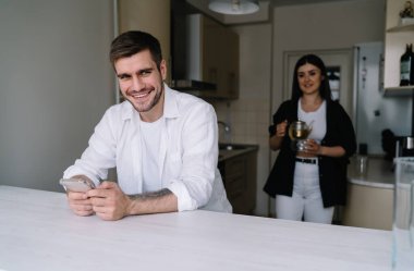 Mutfaktaki masada cep telefonuyla oturmuş sıcak çay ve kupa servisi yapan genç adam gülümsüyor ve başka tarafa bakıyor.