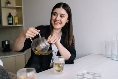 Mutfakta elinde fincanlarla oturmuş cam çaydanlıktan çay dökerken gülümseyen ve mahsulü kesen genç bir kadın.