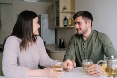Evde romantik vakit geçirirken çay kavanozuyla masaya oturup bardaklara dokunup el ele tutuşan pozitif genç çift.